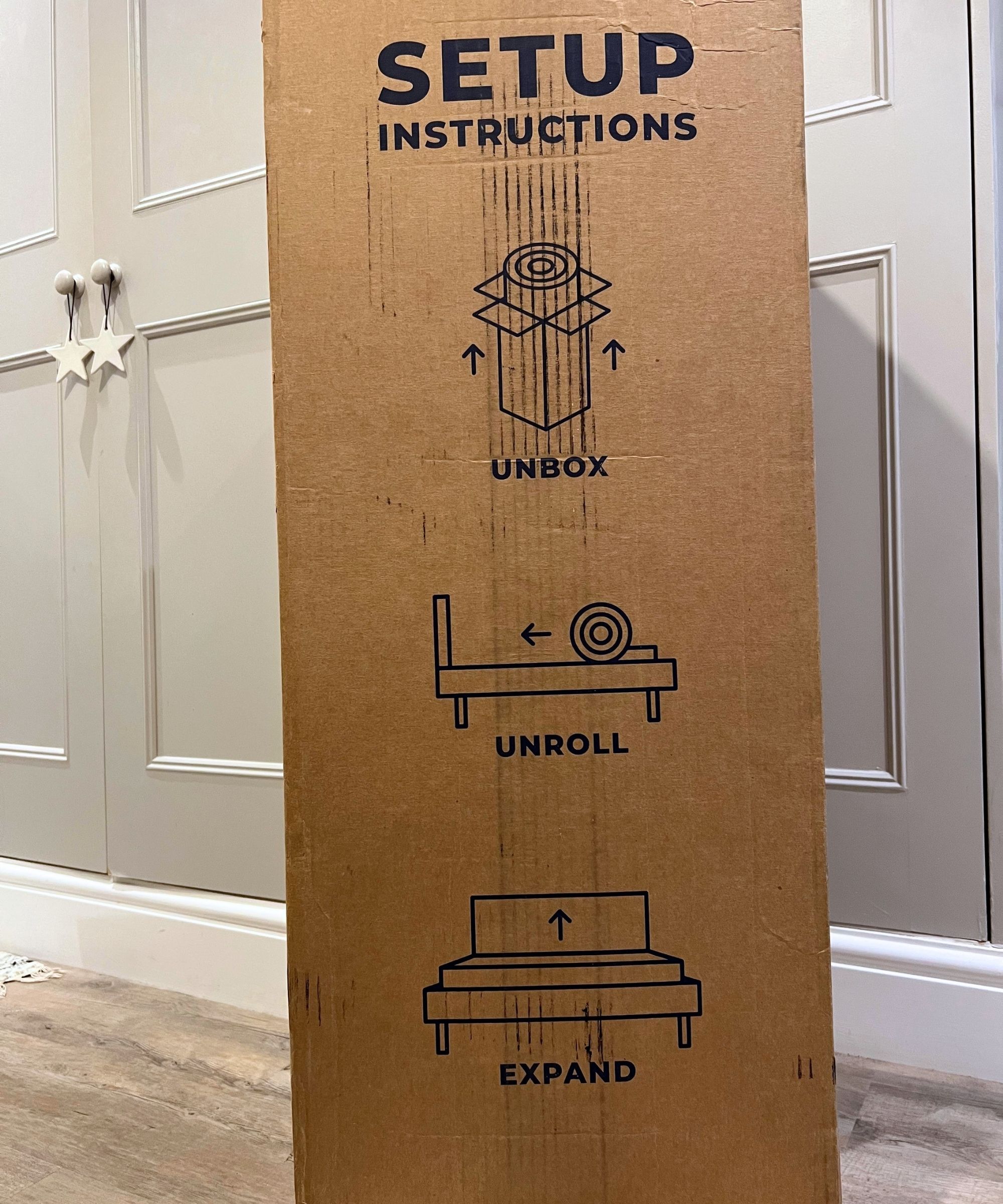 A large cardboard box with unboxing instructions for a mattress topper printed on the side, photographed in front of grey painted cupboards with light wood flooring.