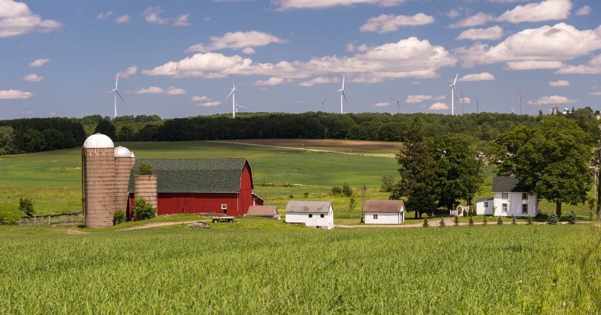 Red barn, silos, and green farm fields under a blue sky, with wind turbines lining a distant treeline.