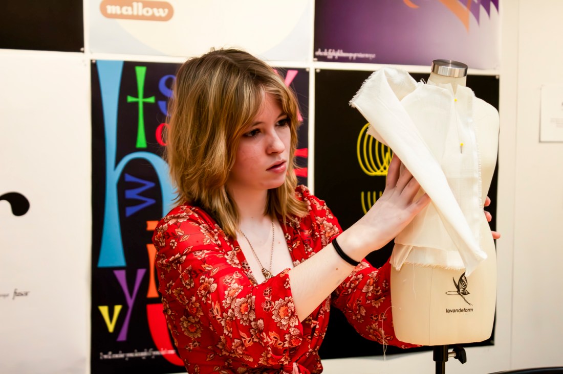 A student holds a piece of clothing they are designing up against a mannequin. 