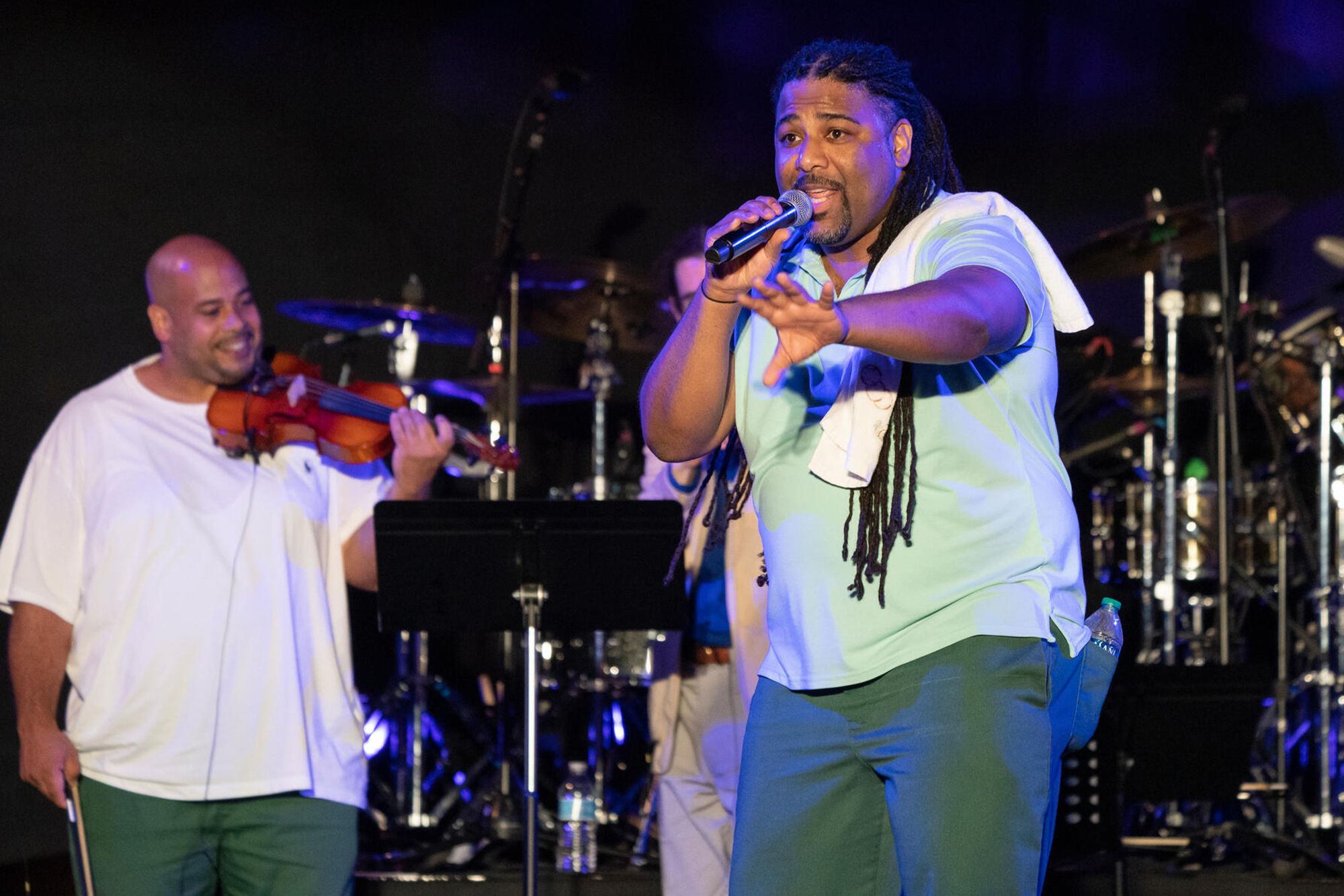 A photo shows a Black man in a pale green T-shirt and dark green pants singing into a microphone. To the left is a man with brown skin holding a violin. 