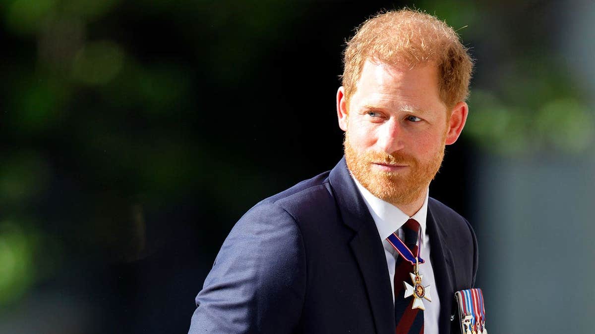 A close-up of Prince Harry in a dark blue suit with medals looking behind him outdoors during a sunny day in London.