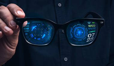 A person holds smart glasses displaying digital interfaces and data, including a clock, date, and various futuristic graphics, projected on the lenses against a dark background.