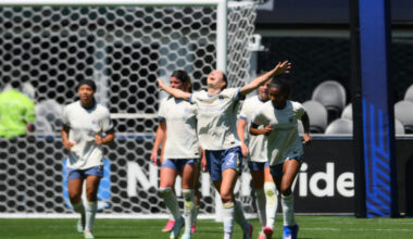 A player in white looks to the sky and raises her arms in celebration. Several other players in white walk behind her.