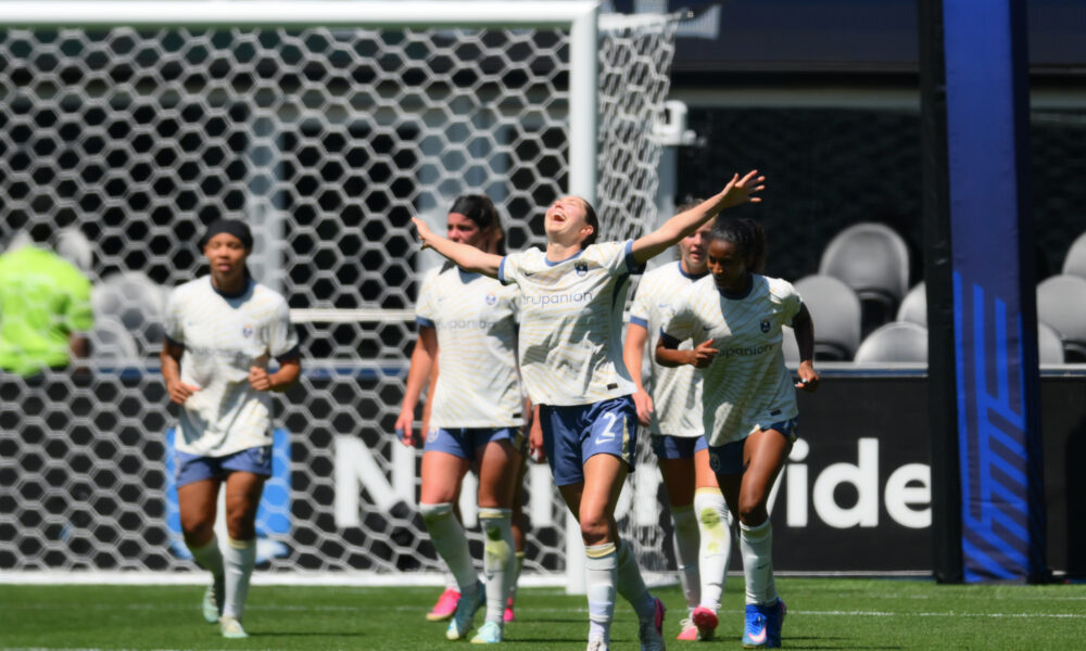A player in white looks to the sky and raises her arms in celebration. Several other players in white walk behind her.