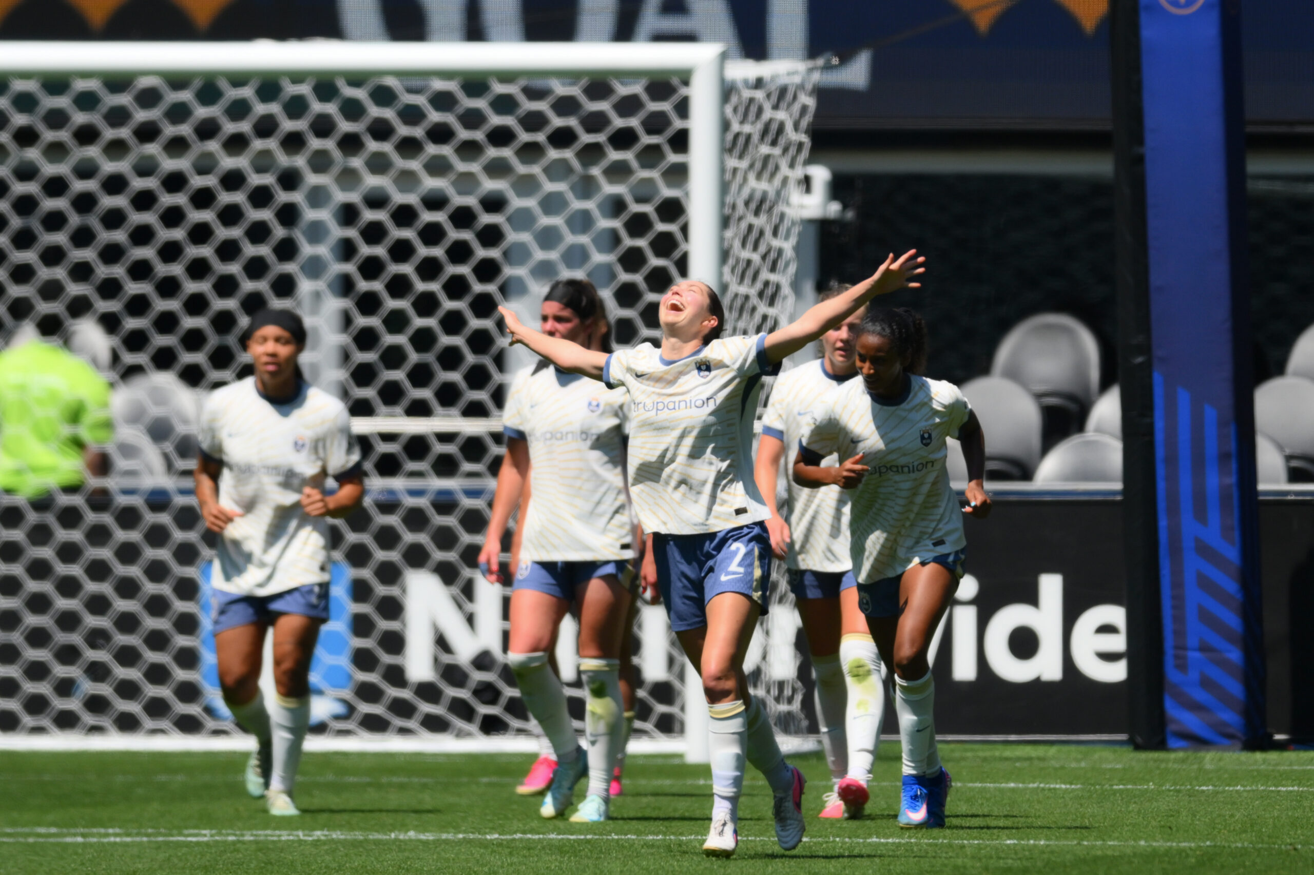 A player in white looks to the sky and raises her arms in celebration. Several other players in white walk behind her.