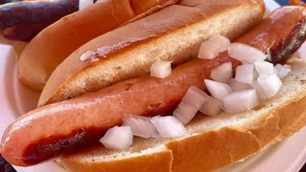 Close-up of a grilled hot dog in a soft bun, topped with diced onions at Heid's of Liverpool. The bun and dog are slightly charred, evoking a classic, casual meal atmosphere.