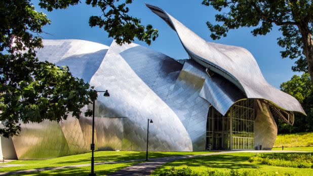 Exterior of the Frank Gehry-designed Fisher Center for the Performing Arts at Bard College, a sculptural, metallic building with curved panels, reflecting light. Surrounded by green grass and trees, exuding a modern, artistic vibe.