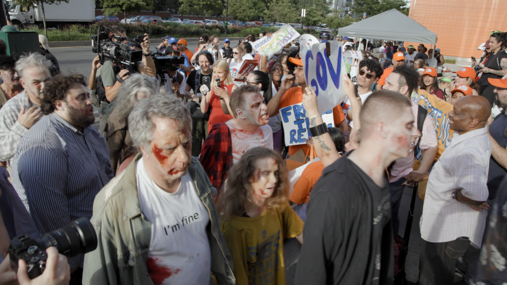 A group of people dressed up like zombies walks past a group of people holding "Freeze the rent" signs.