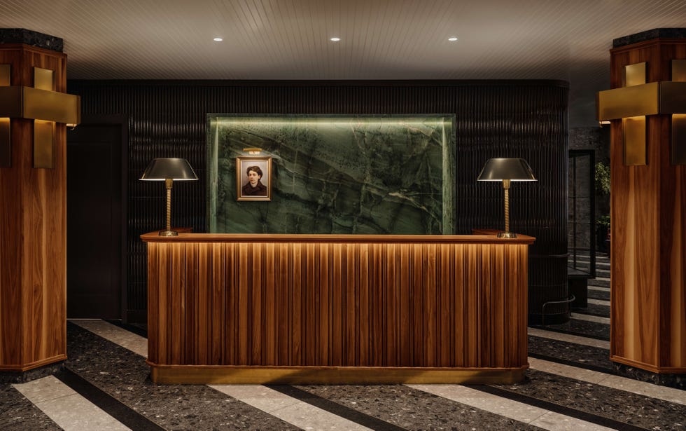 Elegant hotel reception area with wooden counter, green marble backdrop, lamps, and a framed portrait on the wall. Elegant hotel reception area with wooden counter, green marble backdrop, lamps, and a framed portrait on the wall.