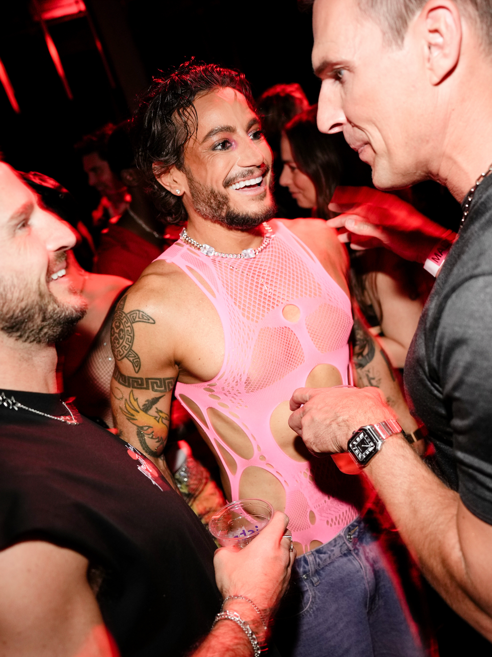 Three people socializing at a lively event, one wearing a bright pink mesh outfit and smiling joyfully.
