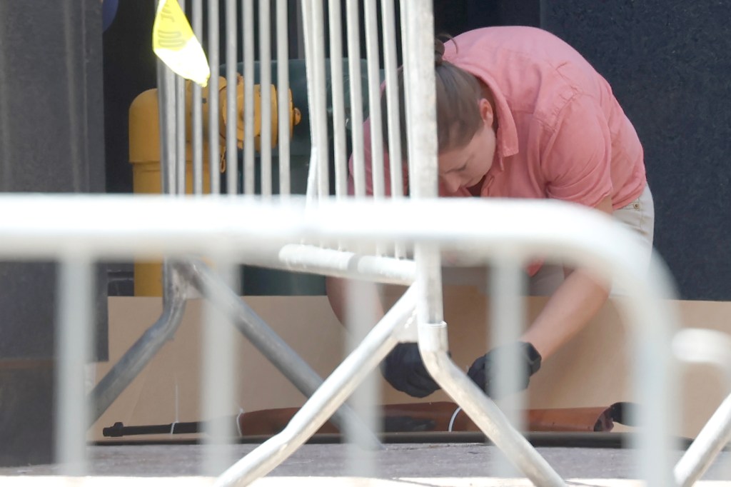 A person in a pink shirt and gloves crouches behind a white barricade to pick up an airsoft rifle.