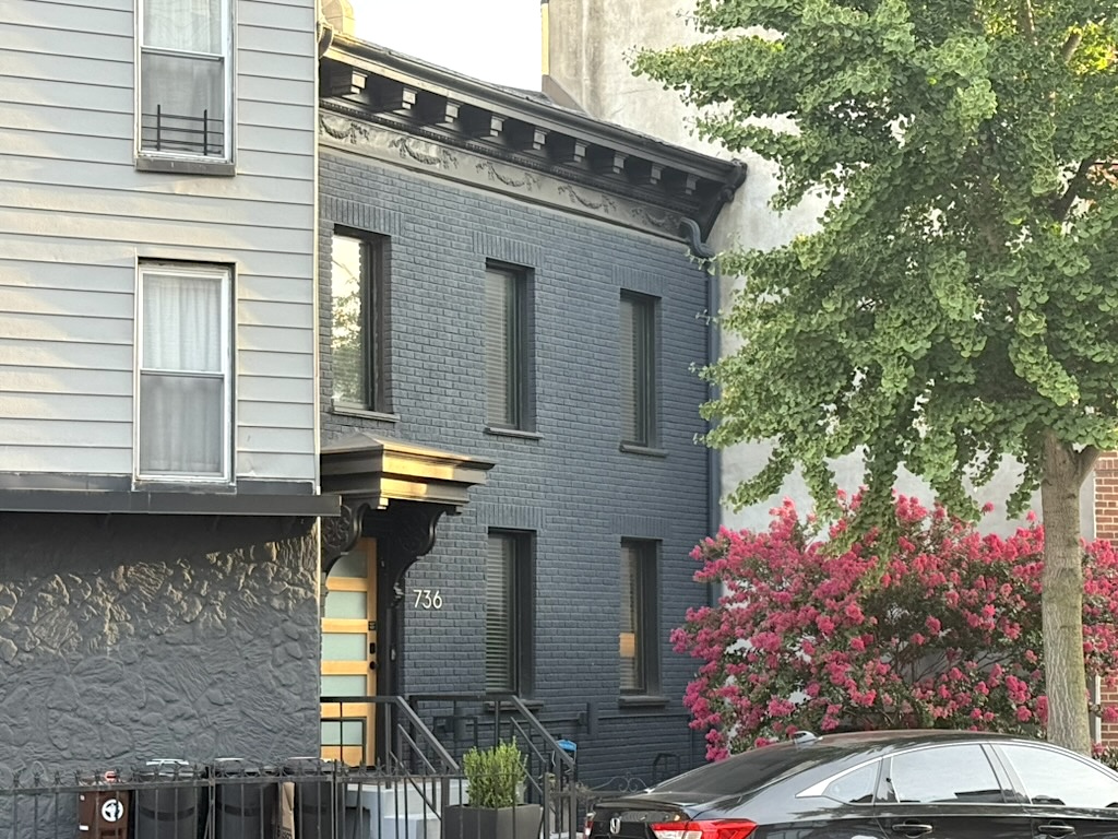 A view of two neighboring buildings, one gray with a textured facade and the other painted black, featuring large windows. In front, there are flowering bushes and a parked black car.