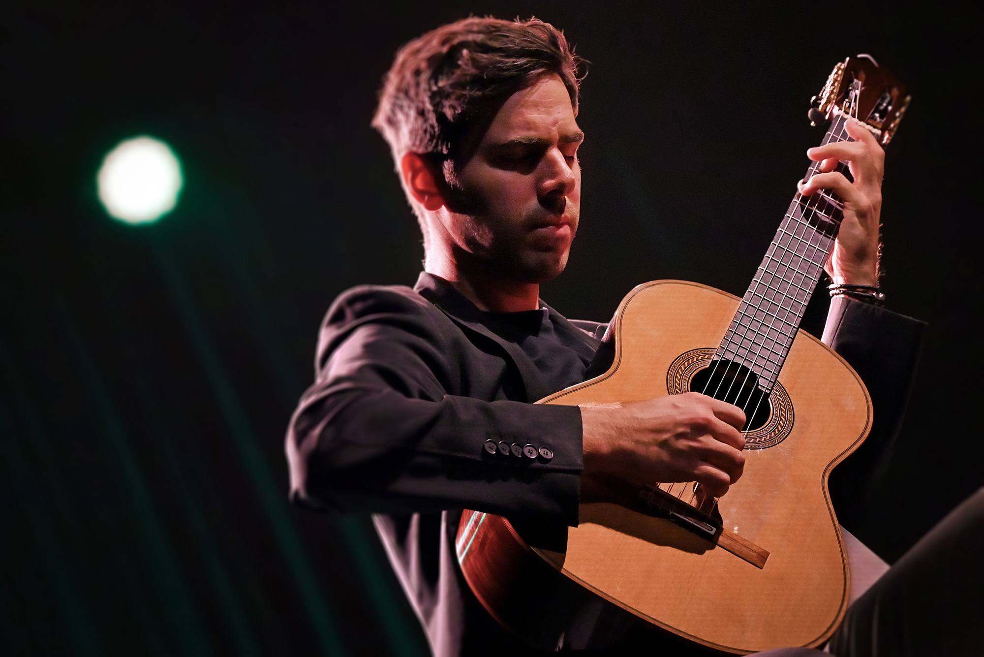 Man playing an acoustic guitar on stage with a dark background and a green light in the distance.