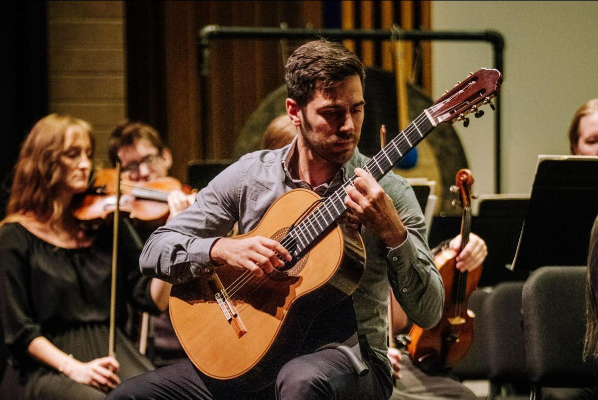 Man playing classical guitar in an orchestra with string musicians in the background.