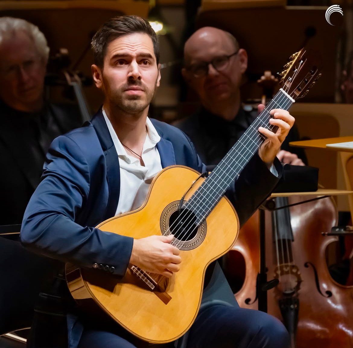 Man playing guitar in an orchestra, with other musicians in the background.