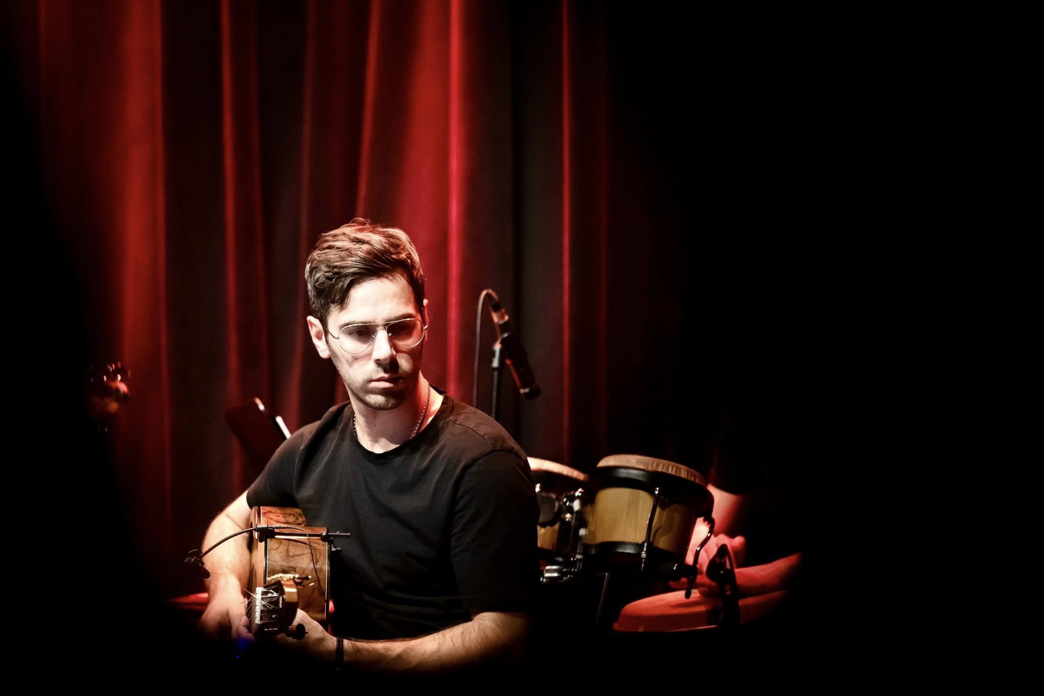 Man with dark hair and beard holding a guitar on stage with red curtains and drums behind him.