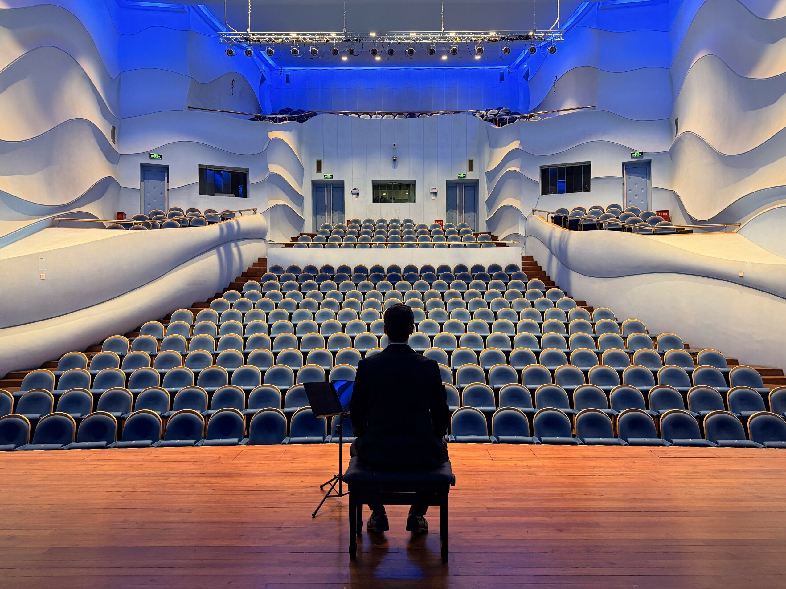 Person sitting on a chair on stage facing an empty auditorium with curved white walls and rows of seats.