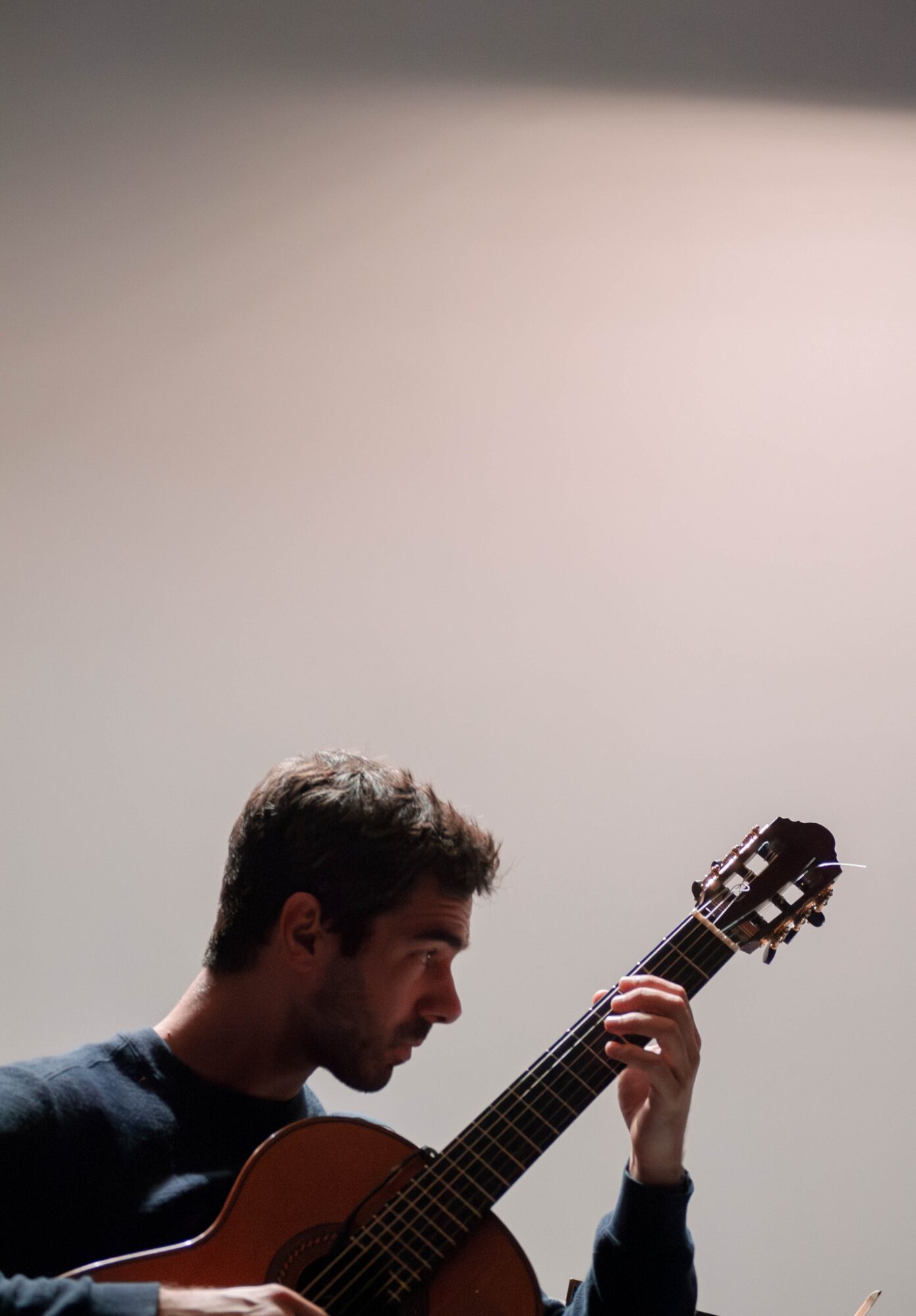 Young man playing guitar against a plain background, focused on his instrument, with short dark hair and a dark shirt.