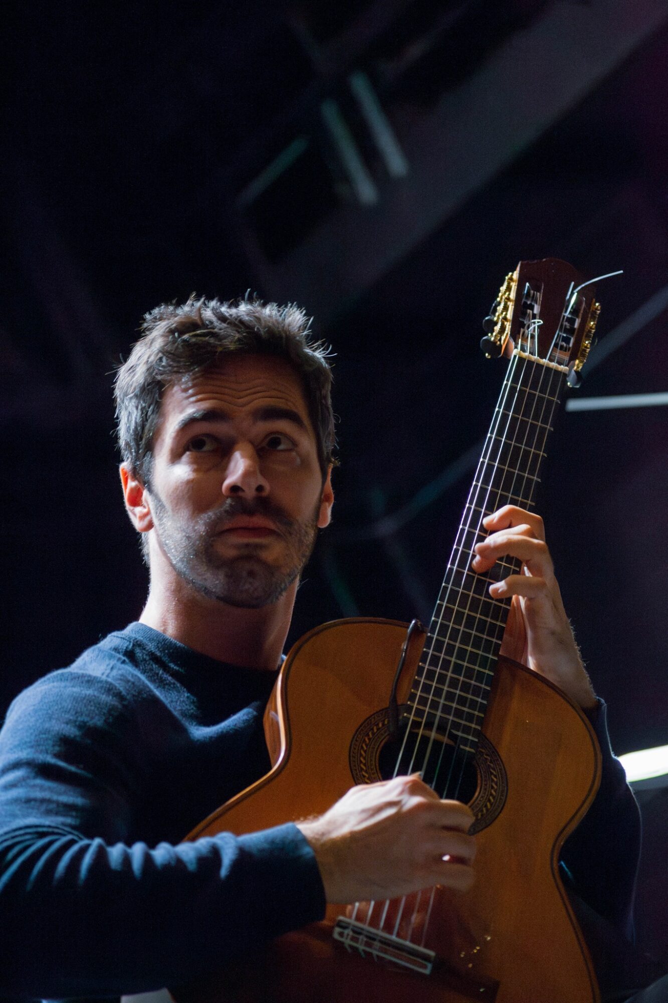 Man holding an acoustic guitar, looking to the side, with dark background and stage lighting.