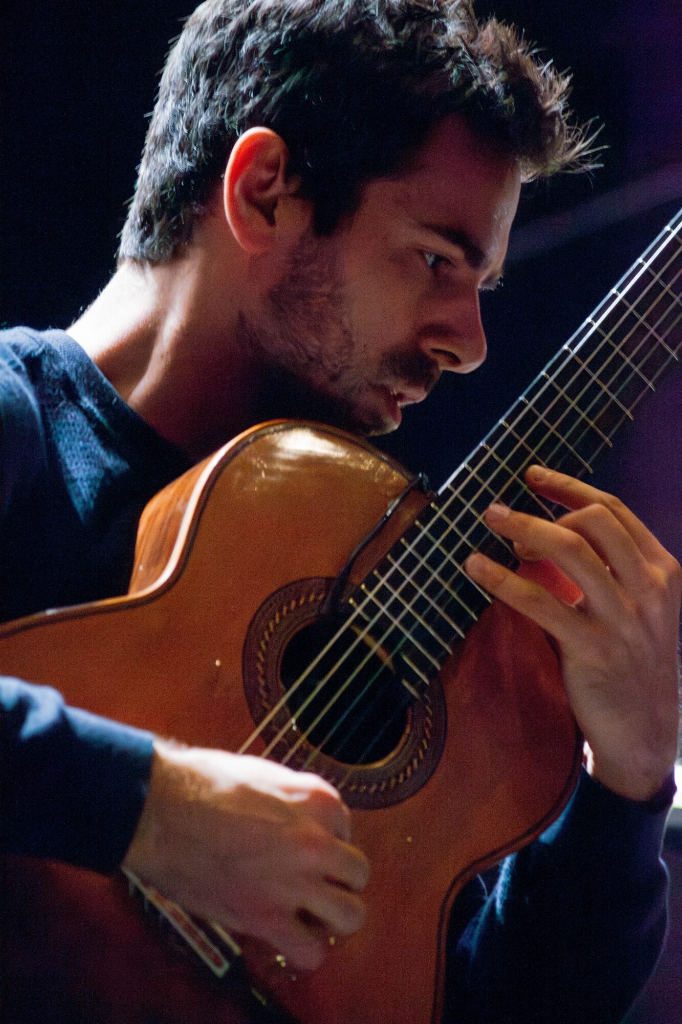 Young man playing an acoustic guitar, focused on the instrument, with dark background.