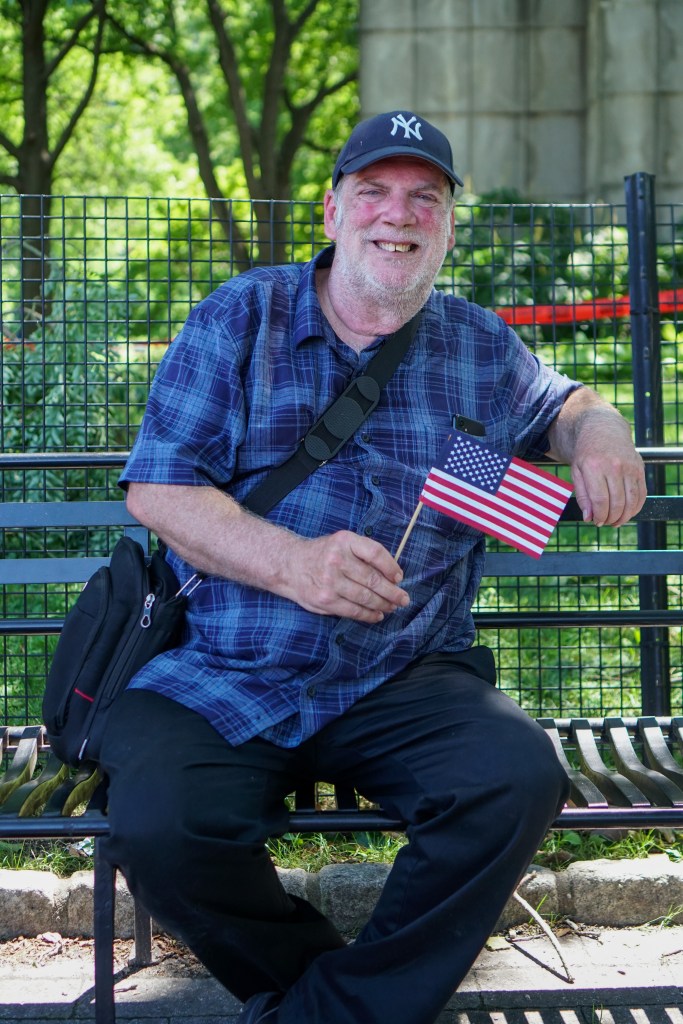 David Bonin, a man in a blue plaid shirt and baseball cap, smiles while holding a small American flag.