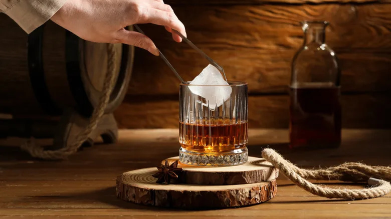 A person adding a large ice cube with tongs to a glass of whiskey