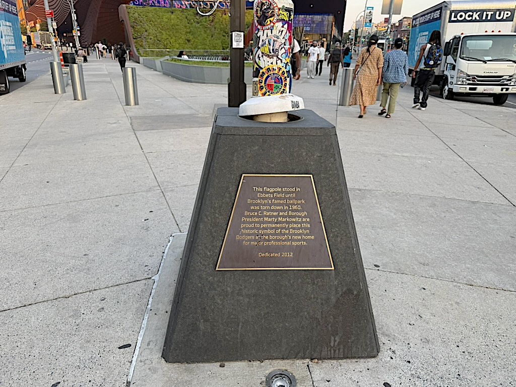 A historical plaque marking the location of the former Ebbets Field in Brooklyn, featuring a flagpole and colorful artwork on top, surrounded by pedestrians and city scenery.