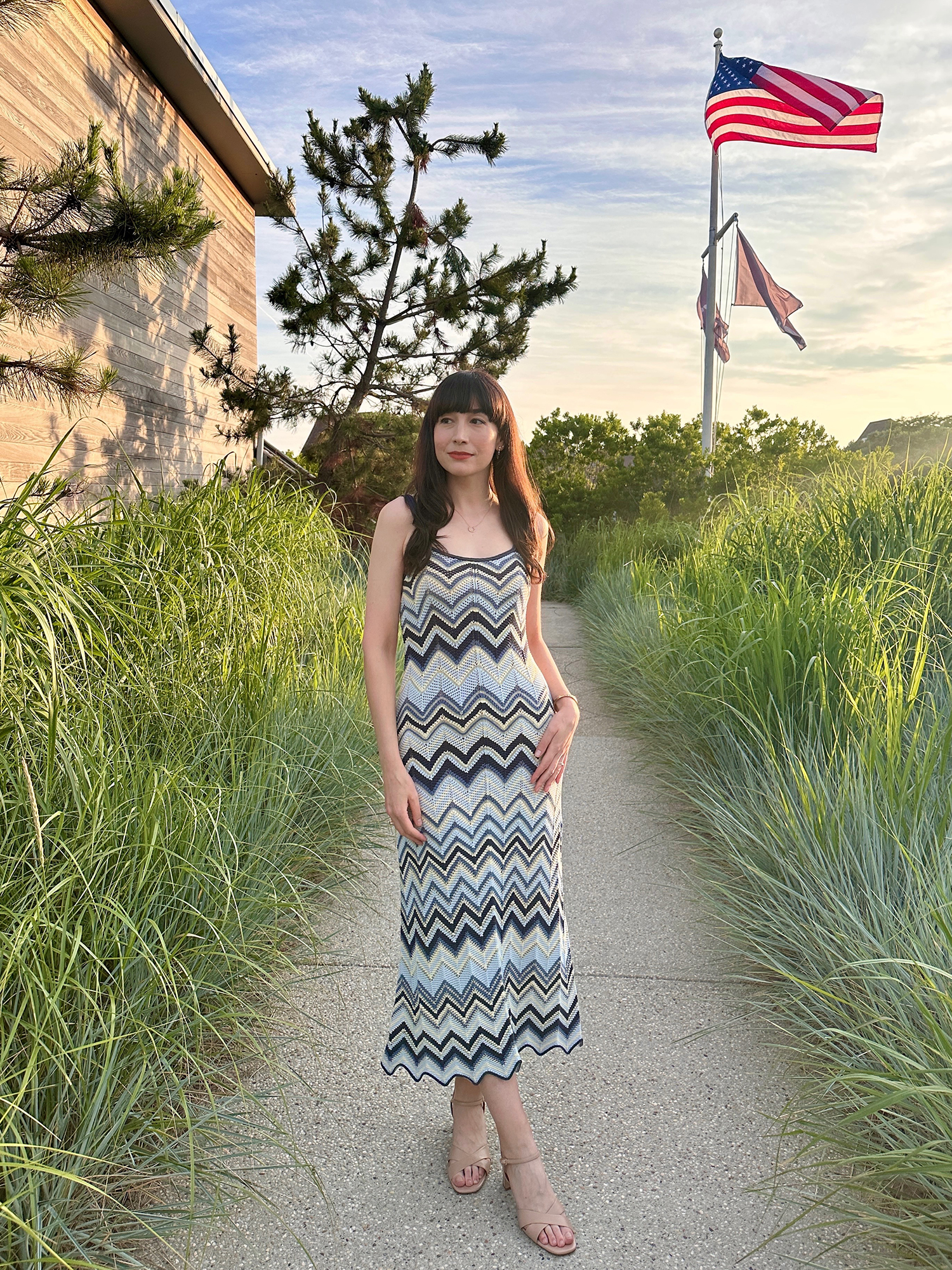 Woman in a zig-zag patterned dress and Paloma sandals standing on a path surrounded by tall grass, with an American flag in the background.