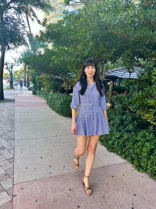 A woman in a blue and white striped shirt dress and brown sandals walks on a Miami sidewalk.