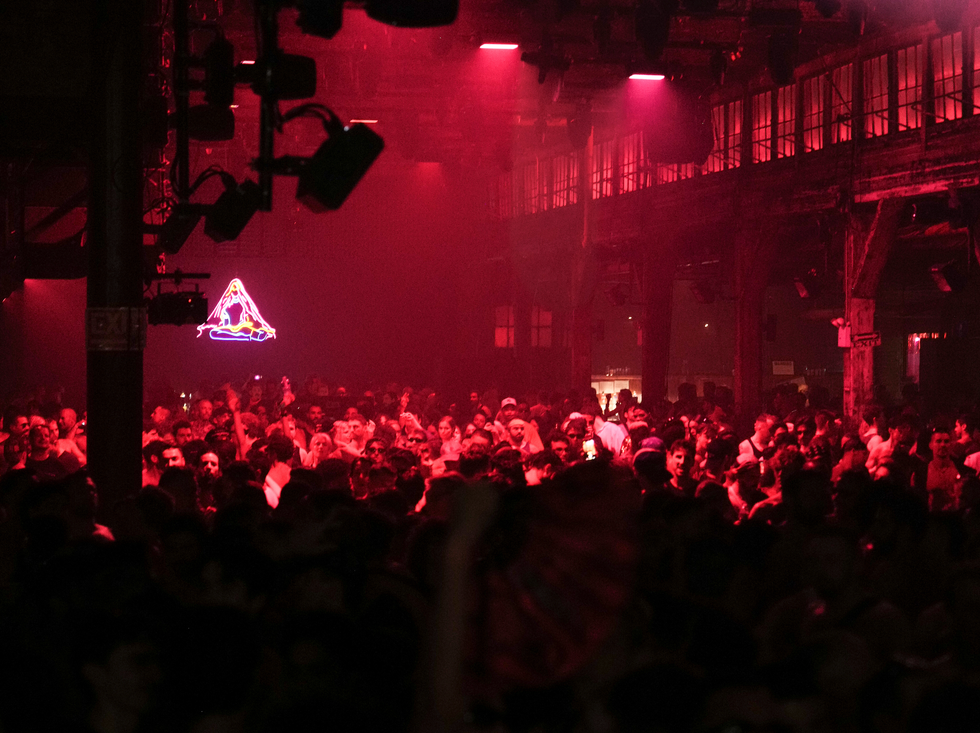 A lively crowd gathered in a dimly lit venue with red lighting and a neon sign in the background.