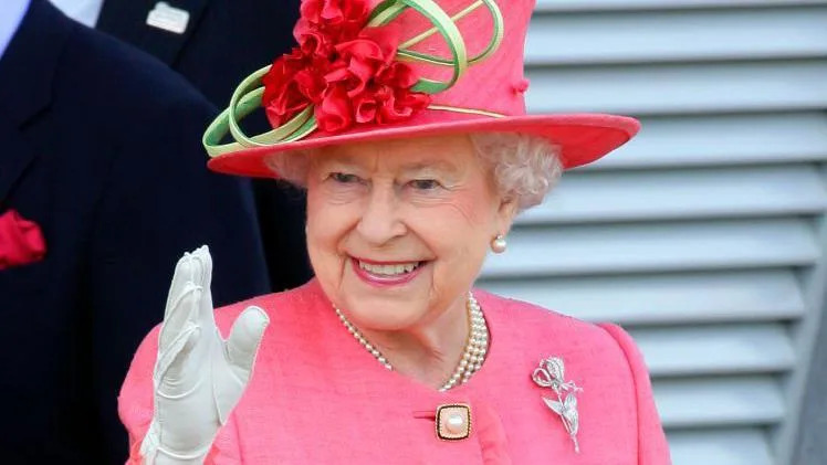 The late Queen Elizabeth II wearing a pink outfit and a hat which has a flower design. One of her hands, in a white glove, can be seen waving in the picture. She is also wearing a silver broach. 