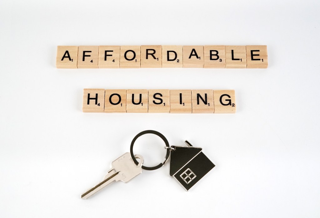 Scrabble tiles spell out "AFFORDABLE HOUSING" next to a house keychain and key.