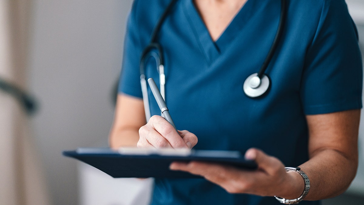 A close-up of a healthcare professional in blue scrubs writing notes on a clipboard