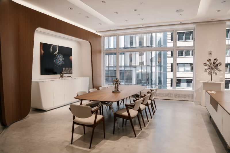 A modern office conference room featuring a large wooden table, light-colored chairs, a white credenza, a wall-mounted display, and large windows with a city view.