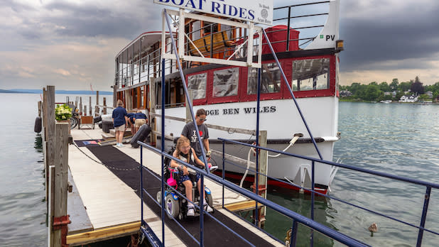 A child in a wheelchair uses a ramp to board a boat named "Judge Ben Wiles" at a dock on Skaneateles Lake.
