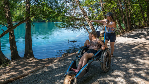 A woman pushes her daughter in a wheelchair along a lakeside path at Green Lakes State Park. The vibrant blue water, surrounded by trees, adds a serene and joyful tone.