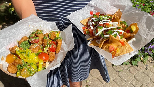 A person holds two dishes outdoors: one with vibrant nachos topped with salsa, jalapeños, and cheese, the other with crispy fried vegetables, garnished with herbs at Taste of Ellicottville.