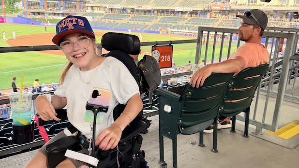 Young woman in a wheelchair, wearing a baseball cap and glasses, smiles at a Syracuse Mets baseball game. A man sits beside her, facing the field. Stadium seats and field are visible.