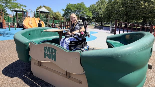 A young girl in a wheelchair smiles while on the "Sway Fun" accessible ride at Wegmans Playground in Onondaga Lake Park. The sunny park features slides and a vibrant, inclusive atmosphere.