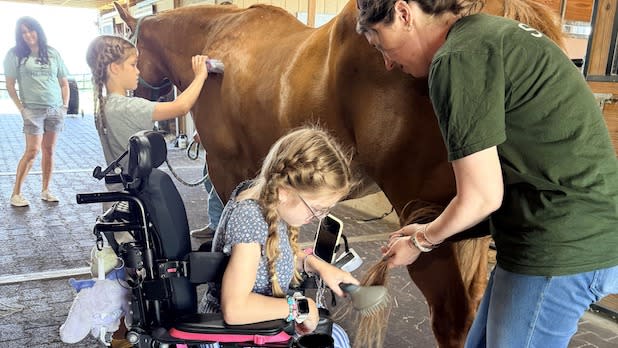 A girl in a wheelchair and a woman interact with a brown horse inside a stable at Root Farm in Sauquoit, NY. Another child brushes the horse's back, while a woman observes nearby. The scene conveys collaboration and care.