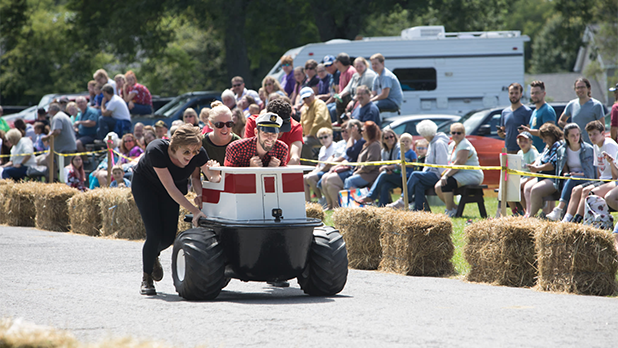 People participate in the Fillmore Days & Bathtub Races, pushing a small, wheeled, boat-like vehicle around a track lined with hay bales. A crowd watches, smiling and cheering.