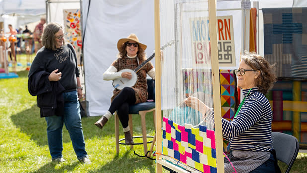 A woman weaves colorful patterns on a loom at the M&T Bank Clothesline Art & Fine Craft Festival . Another woman plays a banjo, creating a lively and creative atmosphere.