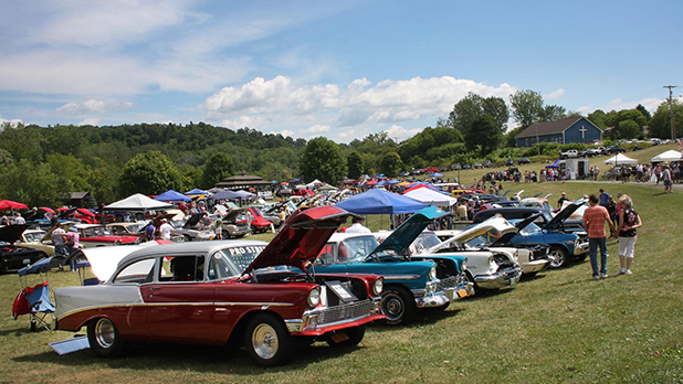 Outdoor car show with classic cars lined up on a grassy field under a sunny sky. People admire vehicles, creating a lively and nostalgic atmosphere at the Ticonderoga Area Car Show.