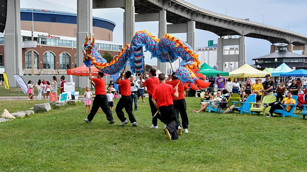 A group performs a colorful dragon dance during the Asian Food and Culture Festival at Buffalo's Canalside. Spectators in bright clothing watch, seated on green grass against a backdrop of event tents and an overpass. The scene is lively and festive.