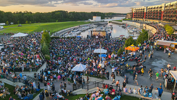 Aerial view of Harbor Jam at the Rivers Casino & Resort in Schenectady, a large, lively outdoor concert by a marina. Crowds fill the area, enjoying live music under colorful tents, with a festive atmosphere.