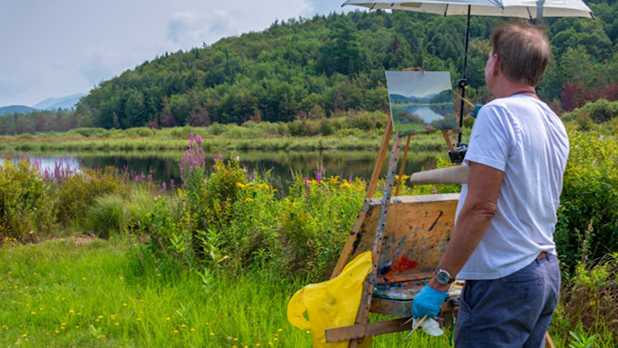 A man in a white shirt paints a scenic landscape on an easel near a calm lake, surrounded by lush greenery and wildflowers, under a sunny sky.