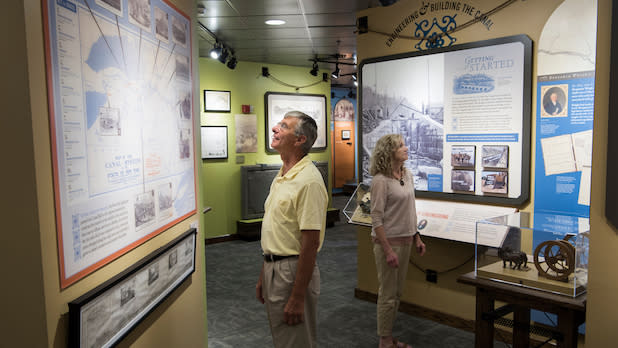 Two people view historical exhibits in the Erie Canal Museum, featuring maps and artifacts. The room is well-lit and has an informative, educational ambiance.