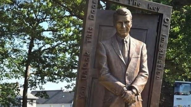 Bronze statue of The Twilight Zone's Rod Serling in a suit holding a door frame with the inscription, "You Unlock This Door with the Key of Imagination," set outdoors in Recreation Park in Binghamton.