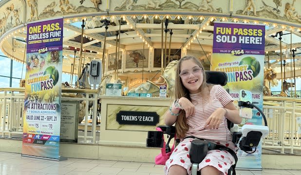 A young girl in a wheelchair smiles in front of the Antique Carousel at Destiny USA. Two colorful "Summer Fun Pass" banners are displayed, creating an inviting, joyful atmosphere.