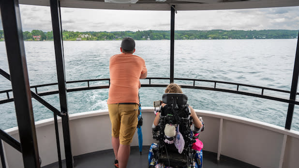 A man stands next to his daughter, who uses a wheelchair, while enjoying a scenic boat ride with Mid-Lakes Navigation on Skaneateles Lake. The landscape features green hills and overcast skies.